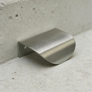 May include: A brushed stainless steel cabinet pull handle with a curved design. The handle is mounted on a concrete surface, showcasing its modern aesthetic. The handle has a small hole on the mounting side.