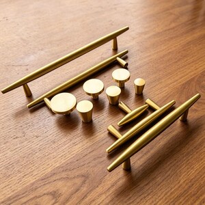 May include: A collection of brushed gold cabinet hardware including two long bar pulls, one shorter bar pull, and several round and tapered knobs. The hardware features a smooth, metallic finish with a consistent golden tone throughout the set.