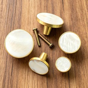 Peut inclure: Un ensemble de quatre boutons de placard circulaires dotés d'une incrustation blanche irisée lisse dans un cadre en métal doré brossé. Deux vis métalliques sont incluses, placées entre les boutons pour montrer les éléments de fixation.