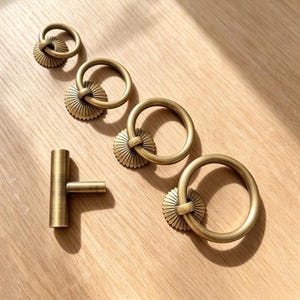Op de afbeelding: Een set messingkleurig beslag bestaande uit drie ronde ringgrepen met geribbelde, zonvormige achterplaten en één cilindervormige T-greep. Elk onderdeel heeft een gladde, metalen afwerking met een consistente gouden kleur.