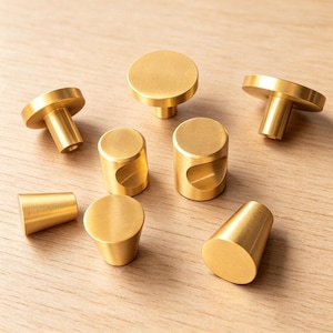 May include: A collection of brushed gold cabinet knobs and pulls in various shapes, including flat circular discs, cylindrical forms with side indentations, and tapered conical designs, all featuring a smooth, metallic finish.