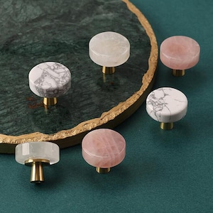 May include: A collection of decorative cabinet knobs. The knobs feature round, polished stone tops in shades of white, pink, and gray, with brass-colored stems. The knobs are arranged on a dark green surface.