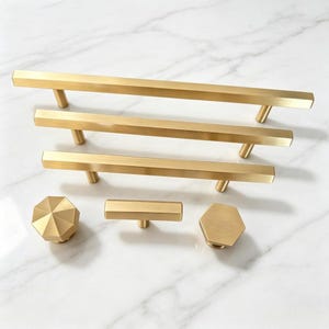 May include: A collection of brushed gold cabinet hardware including three long rectangular bar pulls of varying lengths, one octagonal knob, one small rectangular T-bar knob, and one hexagonal knob, all featuring a smooth metallic finish.