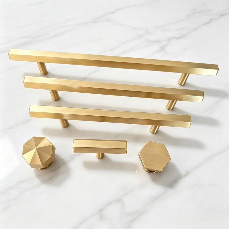 May include: A collection of brushed gold cabinet hardware including three long rectangular bar pulls of varying lengths, one octagonal knob, one small rectangular T-bar knob, and one hexagonal knob, all featuring a smooth metallic finish.