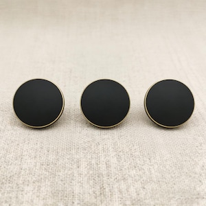 Puede incluir: Tres pomos de armario redondos negros con un borde dorado. Los pomos est&aacute;n dispuestos en l&iacute;nea horizontal sobre un fondo de tela neutra. Los pomos tienen un dise&ntilde;o sencillo y moderno.