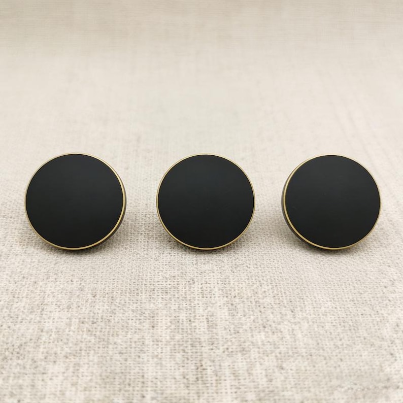 Puede incluir: Tres pomos de armario redondos negros con un borde dorado. Los pomos est&aacute;n dispuestos en l&iacute;nea horizontal sobre un fondo de tela neutra. Los pomos tienen un dise&ntilde;o sencillo y moderno.