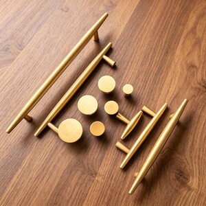 May include: A collection of brushed gold cabinet hardware including two long bar pulls, two shorter bar pulls, and five circular knobs. The hardware features a smooth, metallic finish with tapered ends on the bar pulls and a flat face on the knobs.