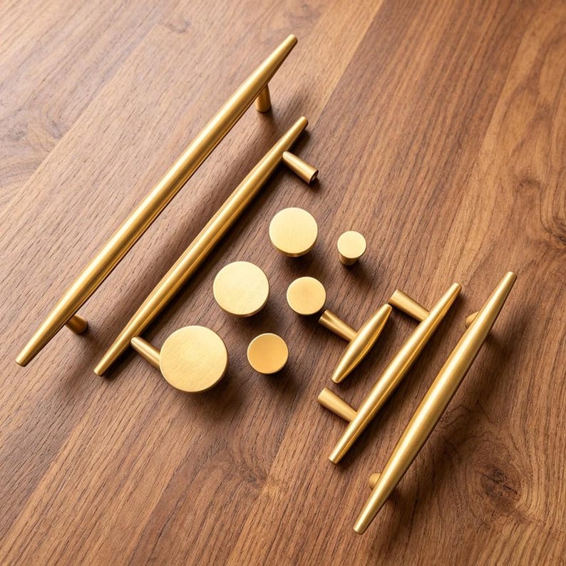 May include: A collection of brushed gold cabinet hardware including two long bar pulls, two shorter bar pulls, and five circular knobs. The hardware features a smooth, metallic finish with tapered ends on the bar pulls and a flat face on the knobs.