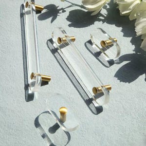 Könnte beinhalten: Vier klare Acryl-Schrankgriffe mit goldenen Metallknöpfen. Die Griffe sind in verschiedenen Größen und Formen, darunter ein langer rechteckiger Griff, ein kürzerer rechteckiger Griff, ein kleiner ovaler Griff und ein größerer ovaler Griff.