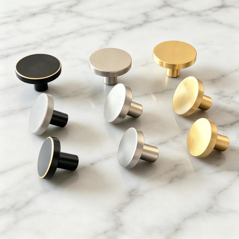May include: A collection of circular cabinet knobs in various finishes including matte black with gold edges, brushed silver, and brushed gold. Each knob features a flat, round face and a cylindrical base, arranged in a group on a white surface.