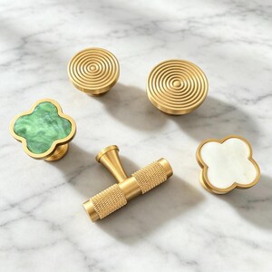 Peut inclure: Un ensemble de cinq boutons et poign&eacute;es de placard aux formes et finitions vari&eacute;es. Deux boutons ronds en laiton pr&eacute;sentent des motifs de cercles concentriques. Une poign&eacute;e en T en laiton a une prise textur&eacute;e. Deux boutons en tr&egrave;fle sont en pierre verte et blanche.