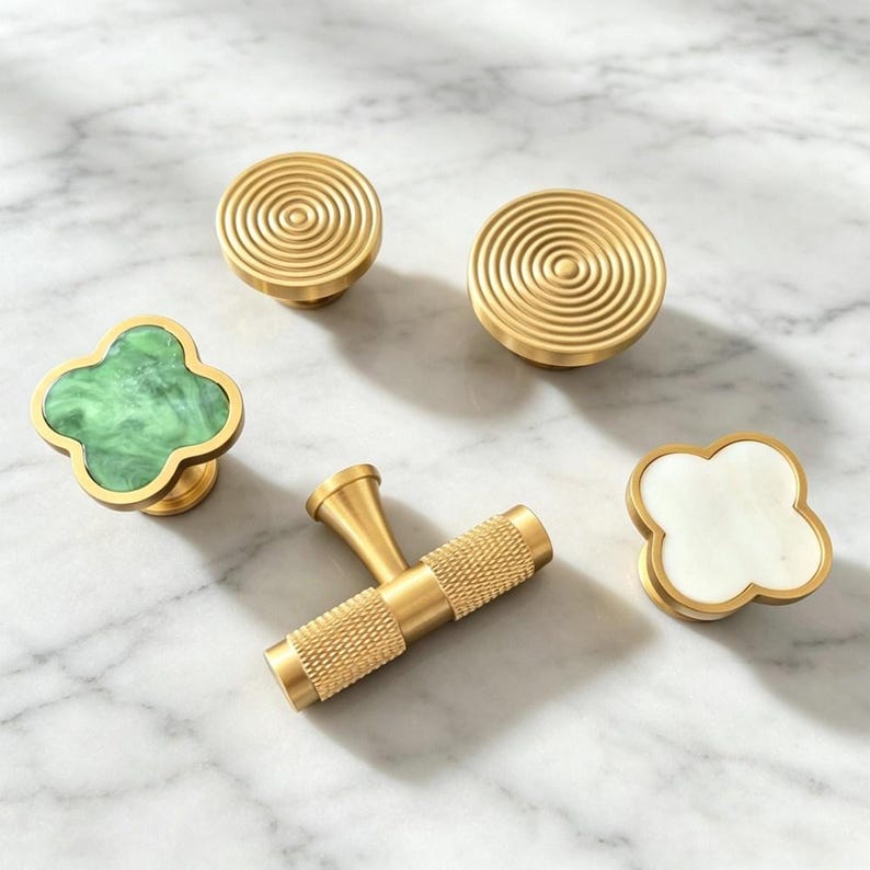 Peut inclure: Un ensemble de cinq boutons et poign&eacute;es de placard aux formes et finitions vari&eacute;es. Deux boutons ronds en laiton pr&eacute;sentent des motifs de cercles concentriques. Une poign&eacute;e en T en laiton a une prise textur&eacute;e. Deux boutons en tr&egrave;fle sont en pierre verte et blanche.
