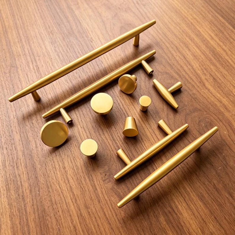 May include: A collection of brushed gold cabinet hardware including long bar pulls, T-shaped pulls, and various circular and conical knobs. The hardware features a smooth, matte metallic finish and is arranged in a group on a wooden surface.