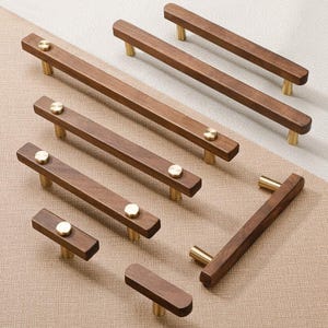 Op de afbeelding: Een verzameling houten kastgrepen in verschillende maten. De grepen zijn rechthoekig met een donkerbruine afwerking en messingkleurige cilindrische palen. Elke greep is bevestigd met een kleine, ronde, messingkleurige schroef.