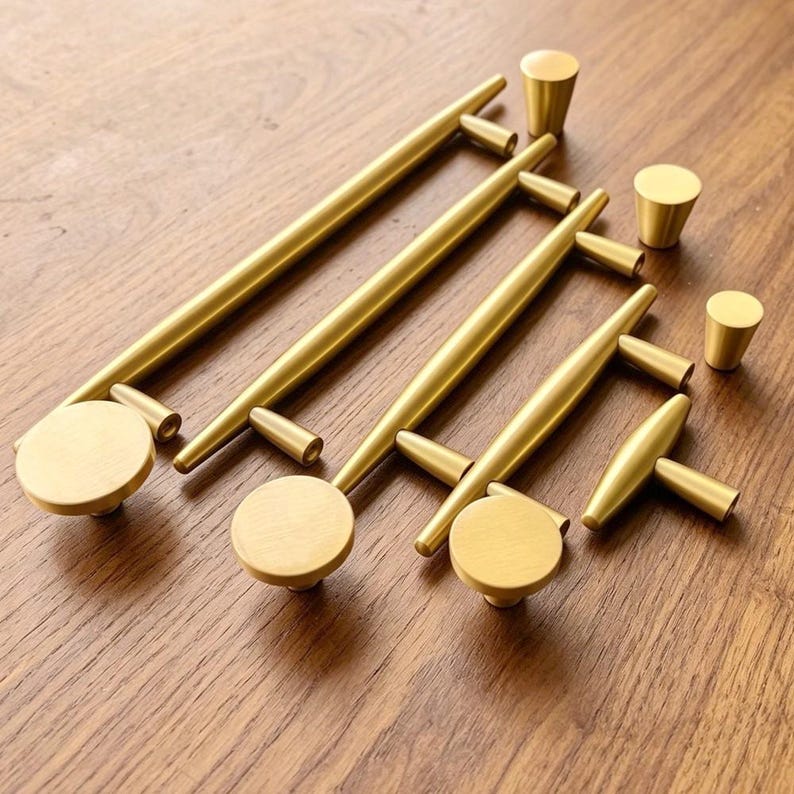 May include: A collection of brushed gold cabinet hardware including three long bar pulls with circular end caps, one T-shaped handle, and three small conical knobs. Each piece features a smooth, matte metallic finish and a cylindrical design.
