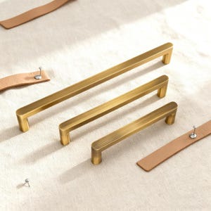 May include: Three brass cabinet pulls of different sizes are displayed. The pulls have a rectangular bar shape with rounded corners and are mounted on short, square posts. Leather straps with metal studs are also visible.