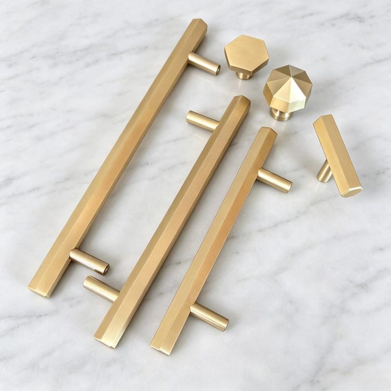 May include: A collection of brushed gold cabinet hardware including three long bar pulls with hexagonal profiles, two faceted hexagonal knobs, and one small T-shaped bar pull. Each piece features a smooth, matte metallic finish and geometric shapes.