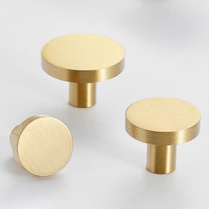 Op de afbeelding: Drie goudkleurige kastknoppen met een geborstelde afwerking. Elke knop heeft een cirkelvormige bovenkant en een cilindrische steel. De knoppen zijn gerangschikt op een wit oppervlak, wat hun eenvoudige, moderne ontwerp laat zien. Geschikt voor laden en kasten.