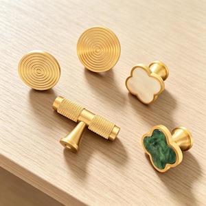 Peut inclure: Un ensemble de cinq boutons et poignées de placard comprenant deux boutons dorés circulaires avec des motifs de cercles concentriques, une poignée dorée en T à surface texturée, un bouton en forme de trèfle avec incrustation blanche et un bouton en forme de trèfle avec incrustation verte marbrée.