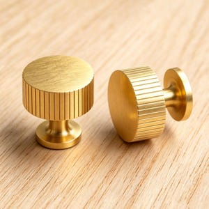 May include: Two gold cabinet knobs. Each knob has a circular top and a cylindrical body with vertical grooves. The knobs are made of metal and are set against a light wood surface. The knobs are suitable for furniture.