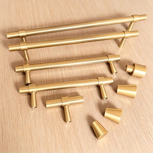 May include: A collection of brushed gold cabinet hardware including three long bar pulls of varying lengths, one T-shaped knob, and four conical knobs. Each piece features a smooth, cylindrical design with a matte metallic finish.
