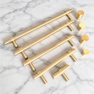 May include: A collection of brushed gold cabinet hardware including four varying lengths of cylindrical bar pulls and four matching cylindrical knobs. Each piece features a smooth, metallic finish with a consistent design and mounting posts.