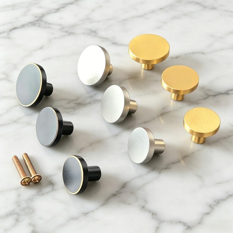 May include: A collection of round cabinet knobs in matte black, brushed silver, and brushed gold finishes. The knobs feature a flat circular face and a cylindrical base. Two metal mounting screws are included with the set of hardware components.