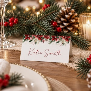 Könnte beinhalten: Eine weiße, gefaltete Tischkarte mit einem gedruckten Blumenrand aus roten Weihnachtssternen, grünen Tannennadeln und kleinen roten Beeren. Der Name Katie Smith ist mittig in einer dunkelroten Schreibschrift aufgedruckt.