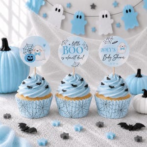May include: Three cupcakes with light blue frosting and black bat sprinkles in light blue cupcake liners with a black spiderweb pattern. Each has a circular topper featuring ghosts, spiderwebs, and text including a little BOO is almost due.