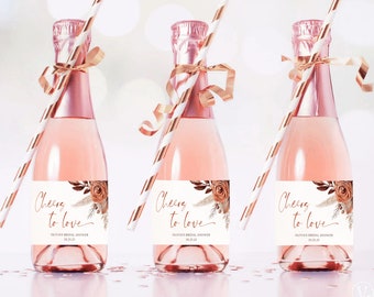May include: Three mini champagne bottles with gold and white striped straws and gold ribbon accents. Each bottle has a white label with a floral design and the text "Cheers to love" and "Olivia's Bridal Shower" with a date.