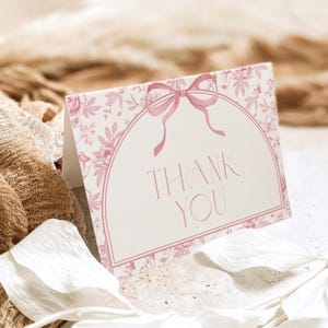 May include: A folded thank you card with a pink floral design and a pink bow. The words "THANK YOU" are printed in a delicate font within a pink arched frame. The card is set against a neutral background.