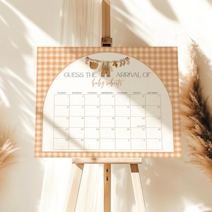 May include: A baby shower calendar on a wooden easel. The calendar has a gingham border and a decorative arch with the text "GUESS THE ARRIVAL OF baby roberts". The calendar grid is for tracking due dates.