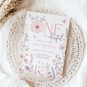 May include: A white rectangular birthday invitation card featuring pink and purple floral illustrations, small butterflies, and the text "isn't she ONEderful" in a mix of script and serif fonts, detailing Ava's 1st birthday event information.