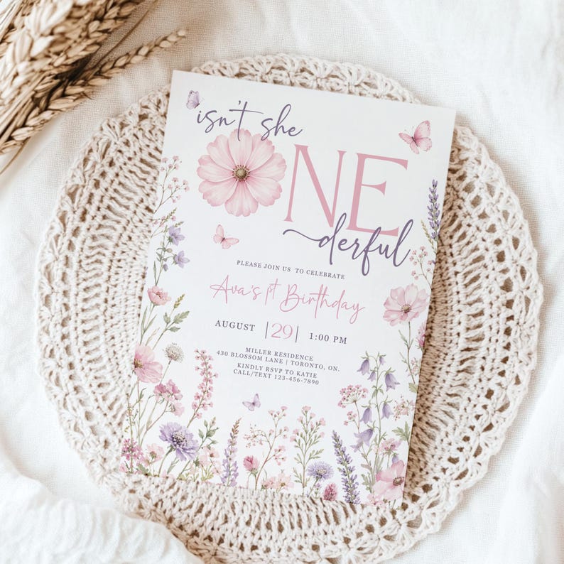 May include: A white rectangular birthday invitation card featuring pink and purple floral illustrations, small butterflies, and the text "isn't she ONEderful" in a mix of script and serif fonts, detailing Ava's 1st birthday event information.