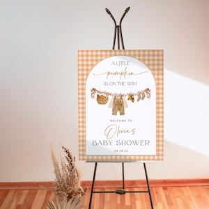 May include: A baby shower welcome sign on an easel. The sign has a tan and white gingham border. The text reads "A little pumpkin is on the way!" with a graphic of baby clothes and a basket. The sign welcomes guests to Olivia's baby shower.