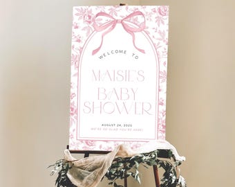 May include: A rectangular welcome sign for a baby shower featuring a pink floral pattern, a large pink bow at the top, and an arched frame. The text reads: WELCOME TO MAISIE'S BABY SHOWER AUGUST 24, 2025 WE'RE SO GLAD YOU'RE HERE! in black and pink.