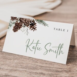 May include: A white place card with a green pine branch and brown pine cones design. The text "TABLE 1" is printed in the top right corner. The name "Katie Smith" is written in cursive in the center of the card.