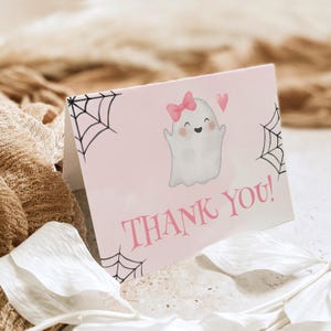 May include: A folded greeting card with a light pink background featuring a white ghost illustration wearing a pink bow, a small pink heart, and black spider web designs in the corners. The text THANK YOU! is printed in bold pink lettering.