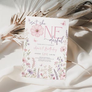 May include: A rectangular birthday invitation card featuring a white background with pink and purple floral illustrations, butterflies, and the text "isn't she ONEderful" in pink and dark purple script and serif fonts, detailing event information.