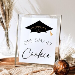 May include: A framed graduation sign with the text "ONE SMART Cookie" in a stylish font. The sign features a black graduation cap with gold accents and the words "Class of 2026". Decorative elements include vases with dried plants.