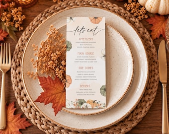 May include: A rectangular paper menu with the text Let's Eat, Appetizers, Main Course, Side Dishes, and Dessert, decorated with watercolor pumpkins and autumn leaves. The menu is centered on a speckled ceramic plate atop a woven round placemat.