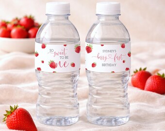 Puede incluir: Dos botellas de agua transparentes con tapas blancas y etiquetas con diseños de fresas. Una etiqueta dice "So sweet to be one", y la otra dice "Sydney's berry first birthday". Fresas frescas están esparcidas alrededor de las botellas.