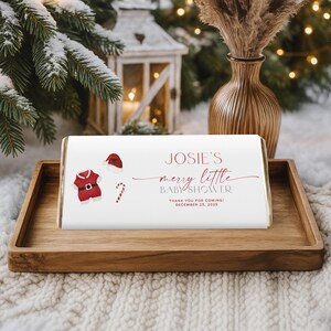 May include: A white candy bar wrapper with red text and Christmas-themed illustrations. The text reads "Josie's merry little baby shower" with a date of December 25, 2025. The wrapper is on a wooden tray.