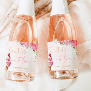Può includere: Due mini bottiglie di champagne con etichette floreali rosa che recitano "Cheers to love" e "Summer's Bridal Shower" con una data. Le bottiglie sono su uno sfondo testurizzato bianco e rosa.