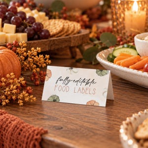 May include: A folded white card with a watercolor pumpkin pattern and the text "fully editable FOOD LABELS" in black and copper-colored lettering. The card is positioned on a wooden surface, featuring a decorative autumn-themed design.
