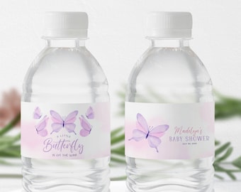 May include: Two clear plastic water bottles with labels. The labels feature a watercolor design of purple butterflies on a pink background. One label says "A Little Butterfly Is On The Way!" The other label says "Madelyn's Baby Shower July 20, 2024".