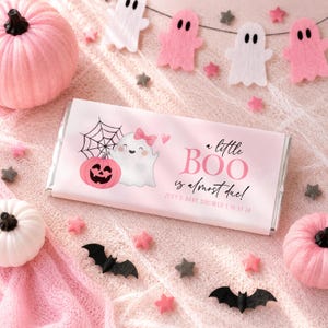 May include: A rectangular chocolate bar wrapper in light pink featuring a white ghost with a pink bow, a pink jack-o'-lantern, and a spiderweb. The text reads: A little BOO is almost due! ZOEY'S BABY SHOWER | 10.27.26 in black and pink font.