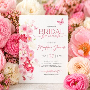 Può includere: Un invito bianco per un brunch nuziale, con composizioni floreali rosa e farfalle. Il testo recita "Bridal Brunch" e include data, ora e luogo. La carta è circondata da fiori rosa.