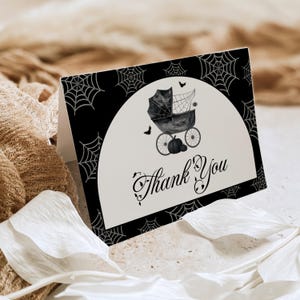 May include: A folded card with a Halloween theme. The card features a black and white design with a baby carriage, bats, and a pumpkin. The words "Thank You" are written in elegant script.