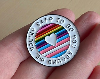 Peut inclure: Épingle ronde en émail avec une bordure blanche et le texte "YOU'RE SAFE TO BE YOU AROUND ME". L'épingle présente un motif à rayures arc-en-ciel avec un cœur blanc au centre.